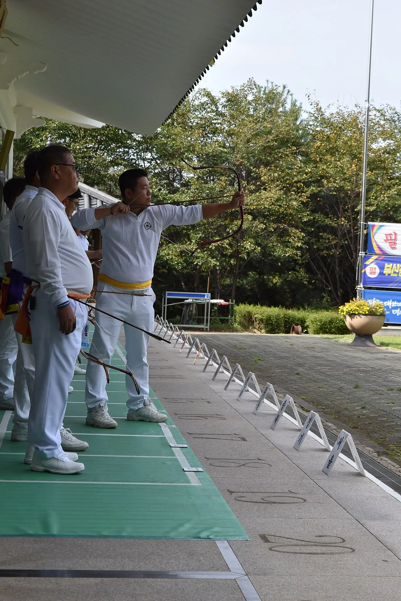 Archer drawing a bow with an arrow nocked on the shooting line, other archers wearing arrow quivers at the waist